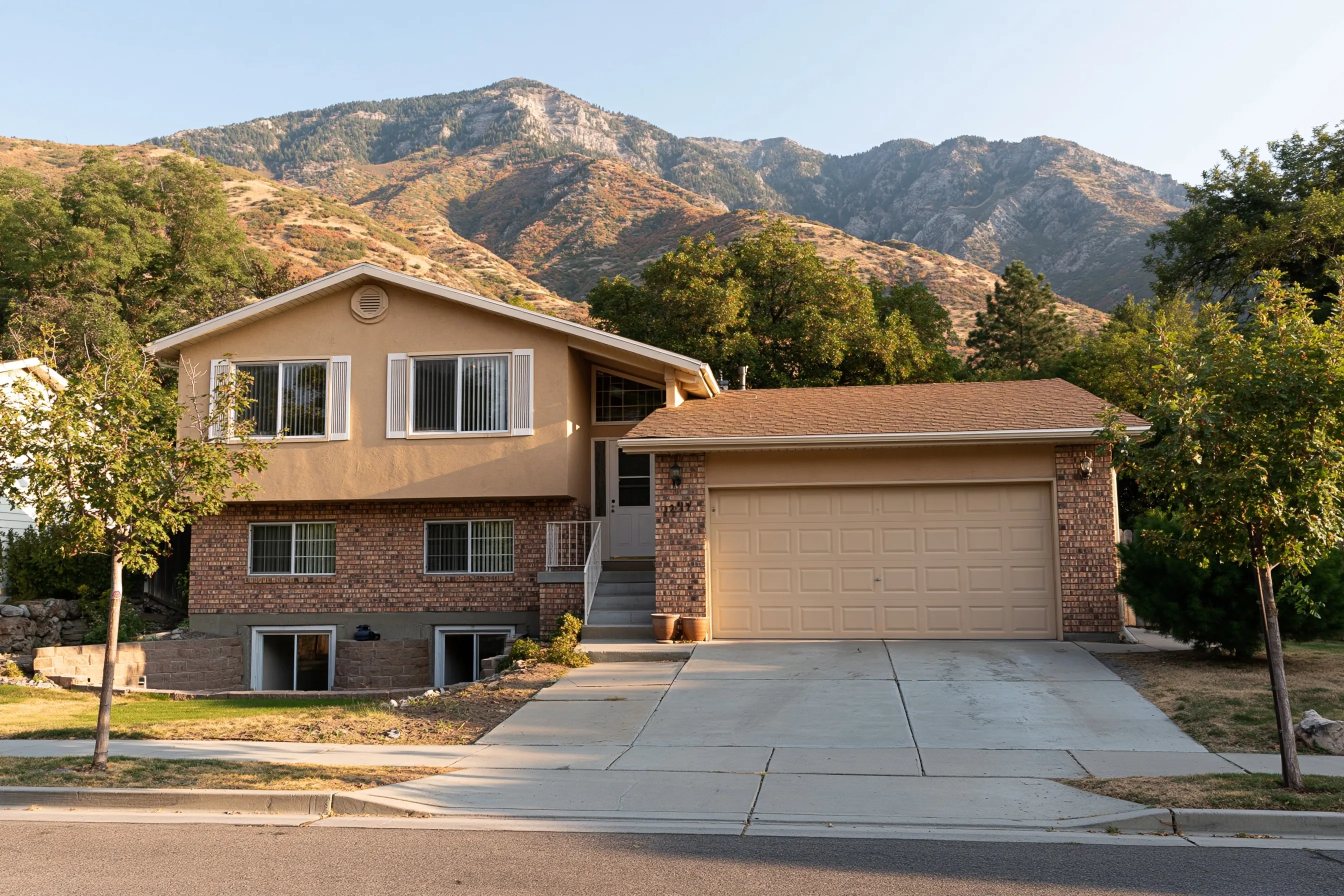 1980s split-level stucco and brick house with a two-car garage, concrete driveway, foundation window wells and scrub oak hills and mountains behind