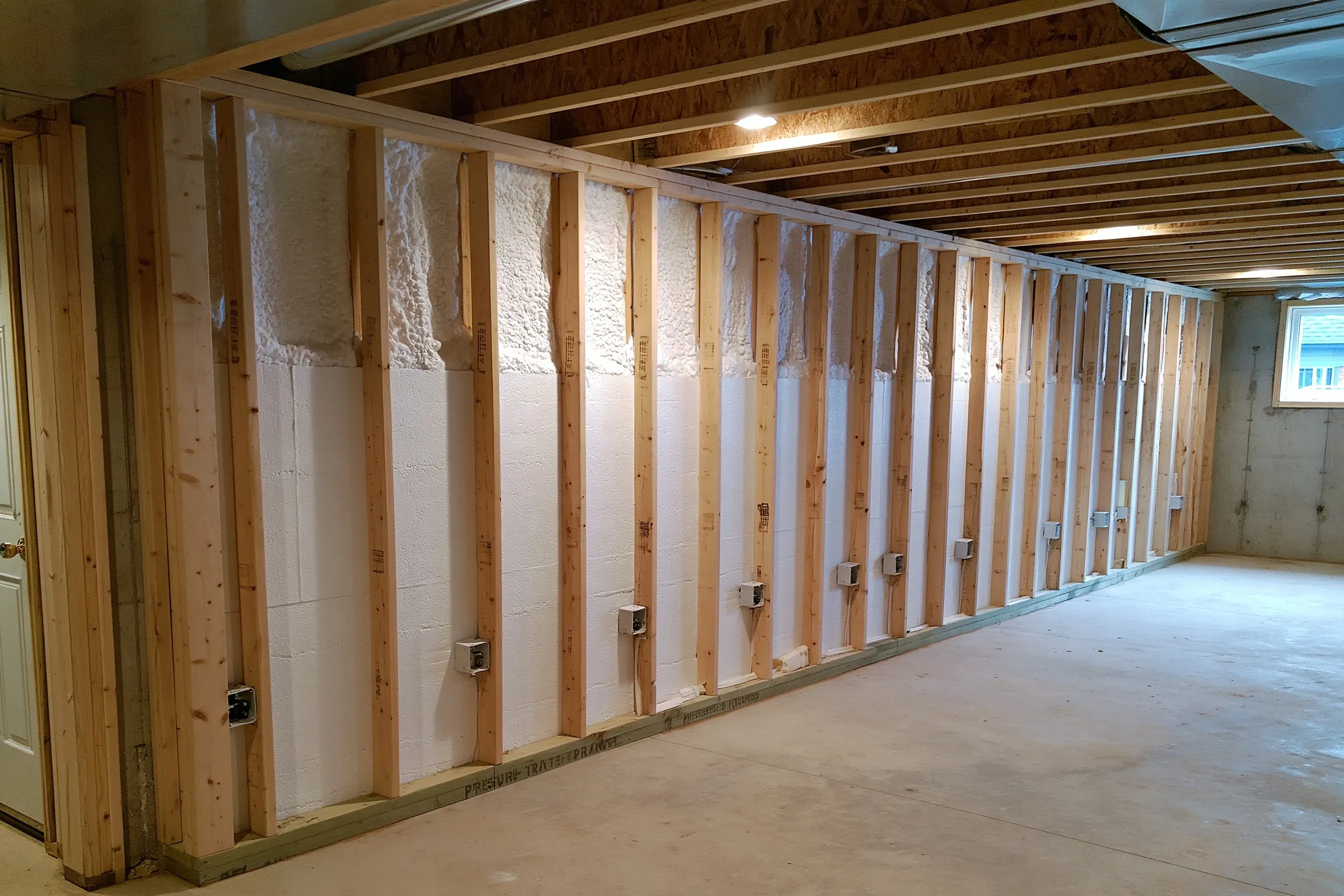 Framed basement wall with spray foam and foam board between the studs, electrical boxes set along the wall, exposed floor joists above and a window at the far end